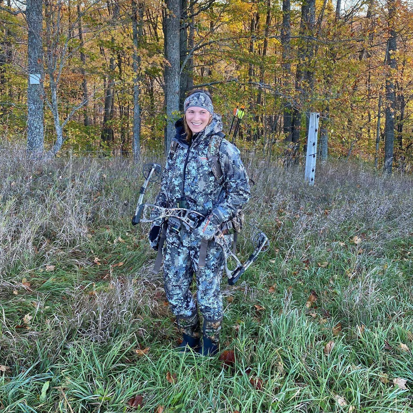 Smiling woman wearing camouflage hunting gear and a bow stands in a grassy field in front of fall foliage.
