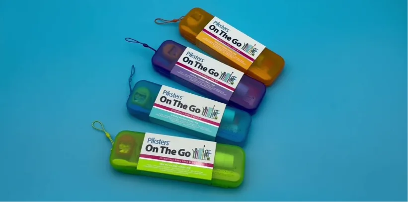 Four colorful “Plackers On The Go” dental floss pick cases stacked diagonally on a bright blue background, each in a different color with a small carrying loop attached.
