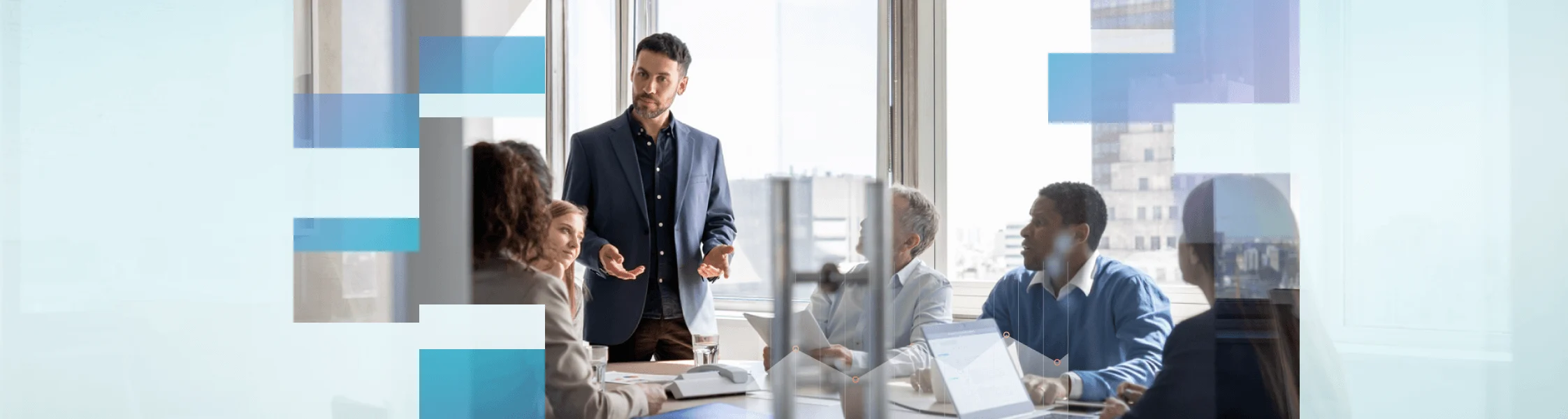 A man in a navy blazer stands and speaks to four colleagues seated at a conference table in a bright office.