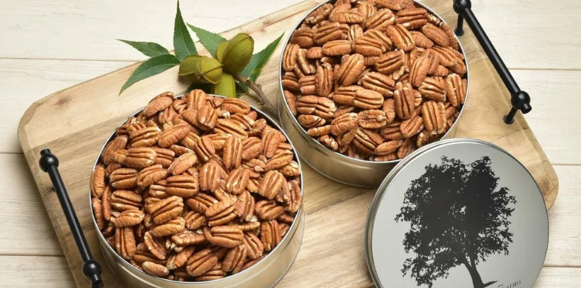 Two metal tins filled with pecans on a wooden tray, with a pecan branch and a tin lid featuring a tree silhouette nearby.