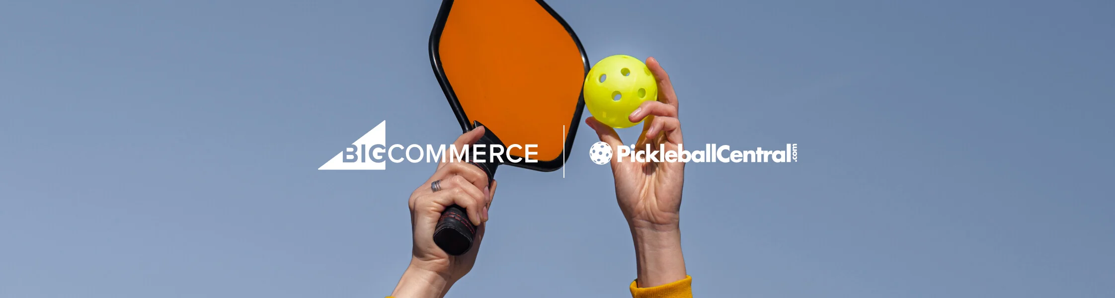 Hands holding a pickleball paddle and ball, with a sky background, and logos for BigCommerce and PickleballCentral.