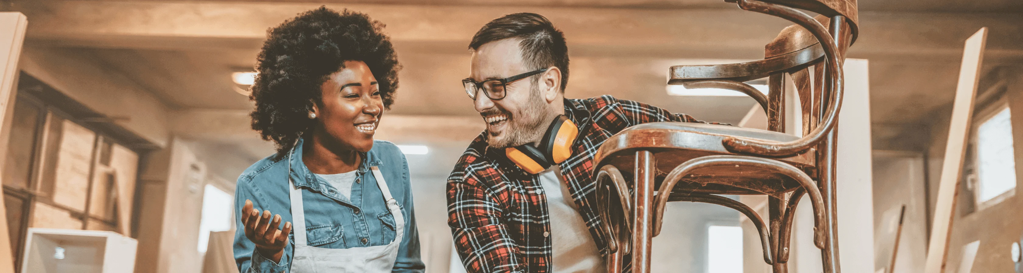 Two smiling woodworkers, a woman and a man, discuss a wooden chair in a bright workshop.