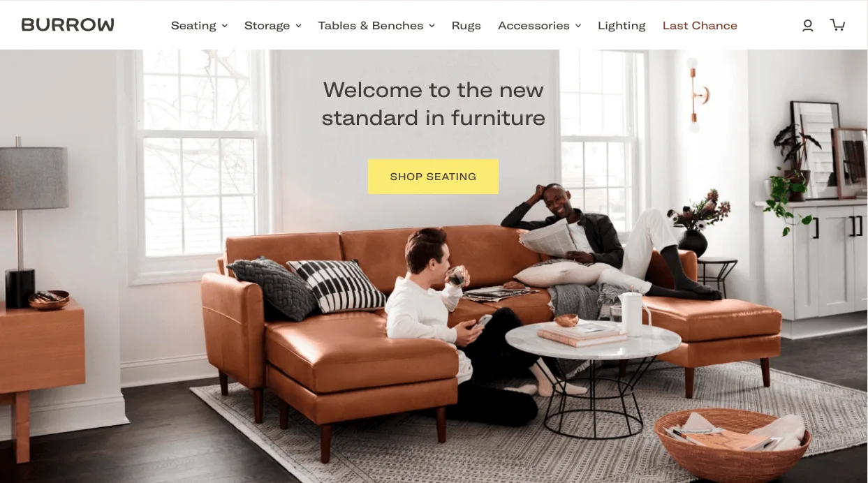 Two men relax in a modern living room on a tan sectional sofa, one reading and one drinking coffee, with bright natural light.