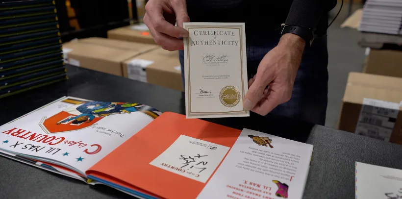 A person holds a Certificate of Authenticity above an open illustrated book on a table with boxes in the background.