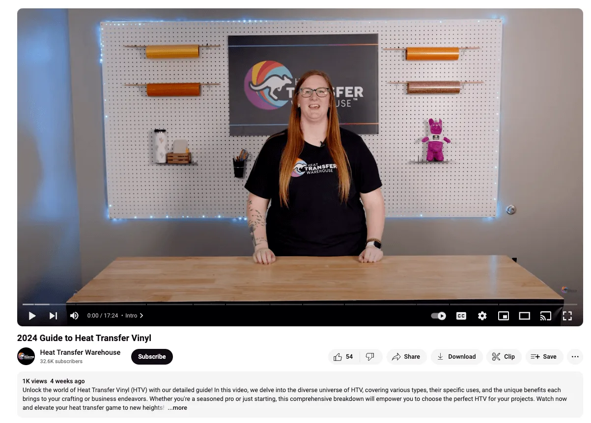 A person with long red hair stands behind a wooden table, smiling, with craft supplies and rolls of vinyl on a pegboard wall.