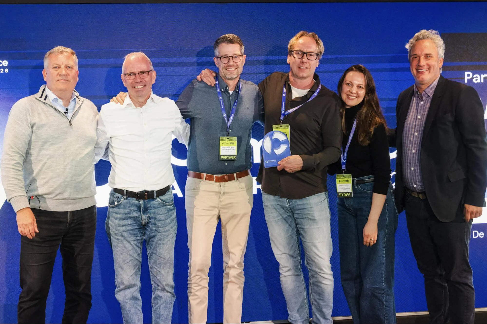 DNZ wins Partner of the Year at the Commerce Awards EMEA 2026. Six people wearing conference badges pose on stage; one holds a blue award trophy against a blue backdrop.