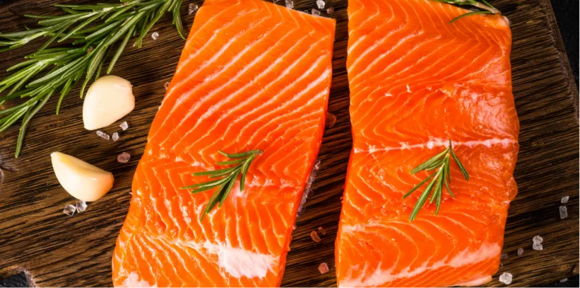 Two raw salmon fillets with rosemary sprigs on a wooden cutting board, accompanied by garlic cloves and peppercorns.