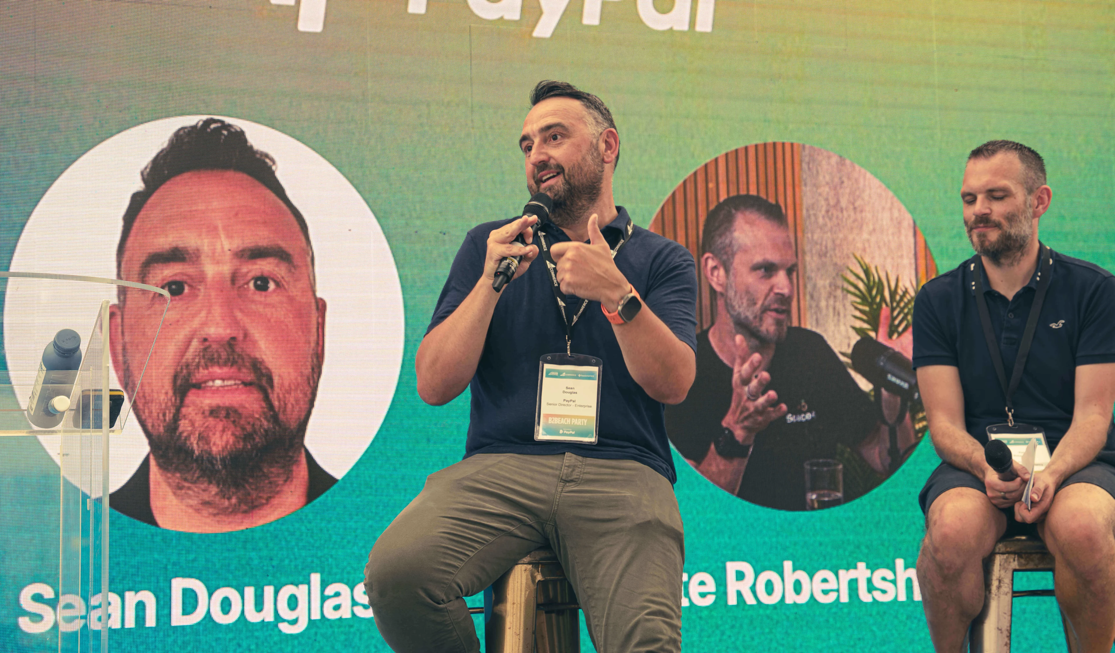 Sean Douglas from PayPal speaking on a panel, giving a thumbs up, with a large headshot of him projected on the screen behind him.