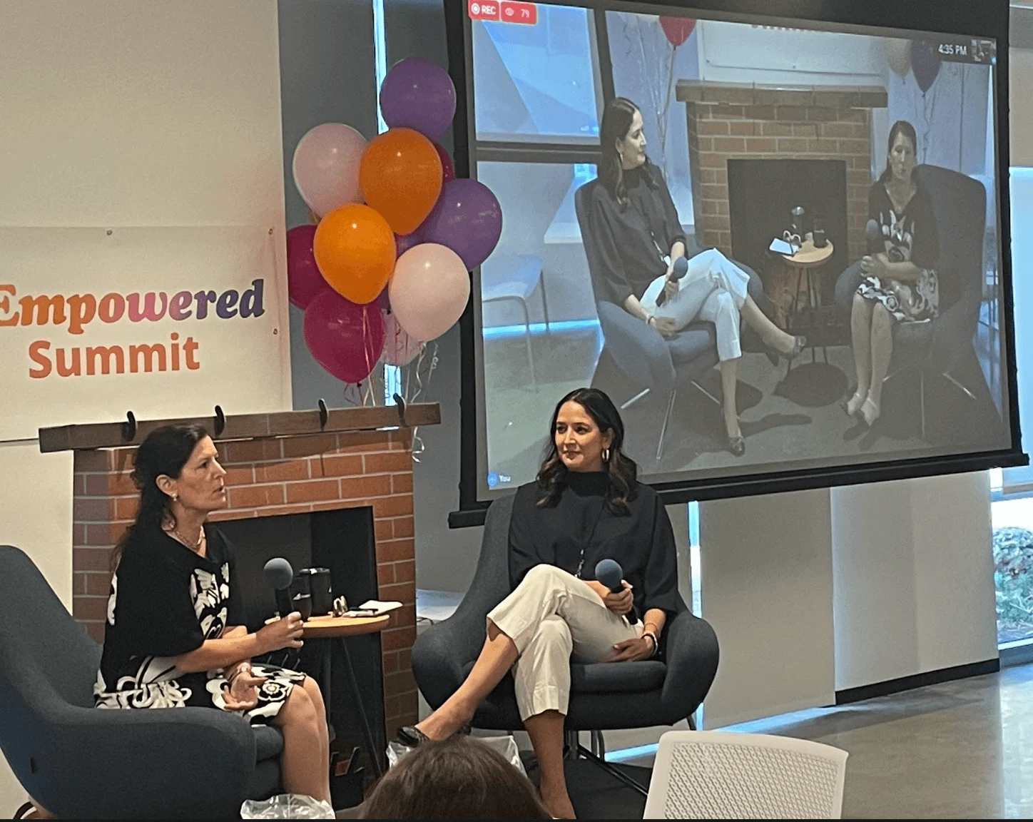 Two women speak at the "Empowered Summit" event, seated in front of a brick fireplace with balloons and a video screen displaying the remote speaker.