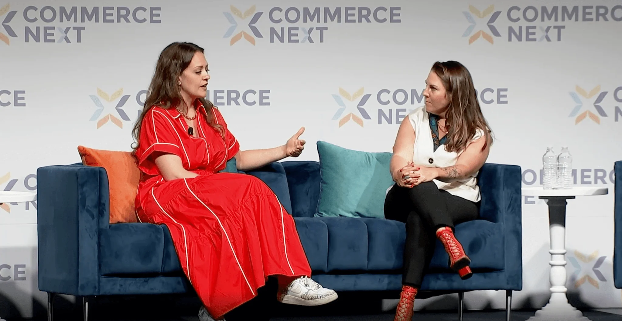 Two women in conversation on a blue sofa at Commerce Next event, one in red dress, one in white top with black pants.