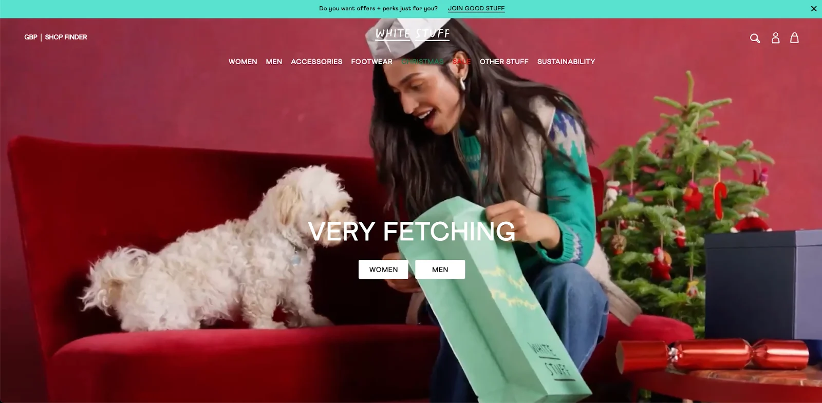 Woman in a green sweater showing a gift bag to a white dog on a red sofa, with a decorated Christmas tree nearby.
