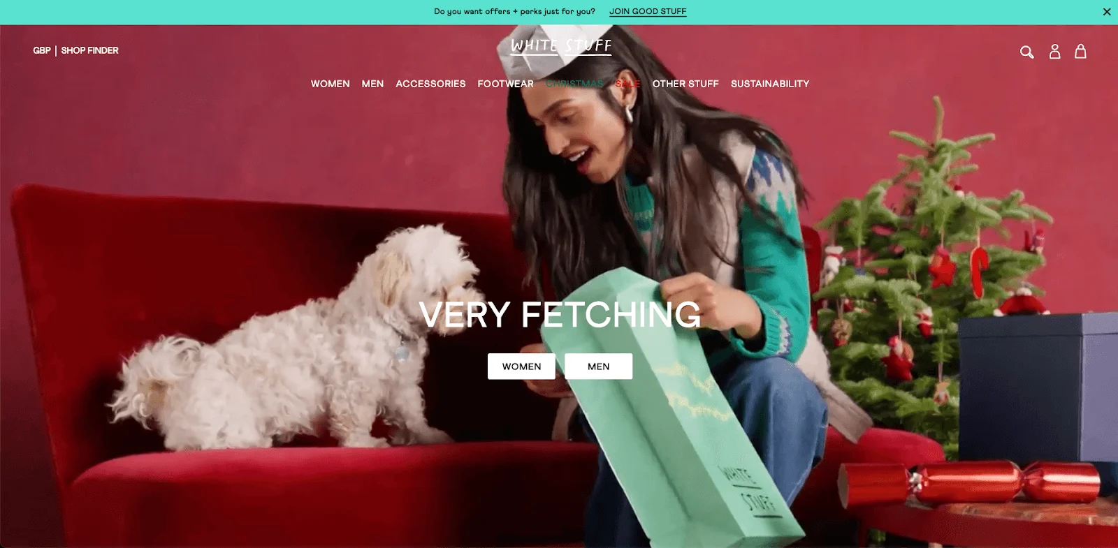 Woman in a green sweater showing a gift bag to a white dog on a red sofa, with a decorated Christmas tree nearby.