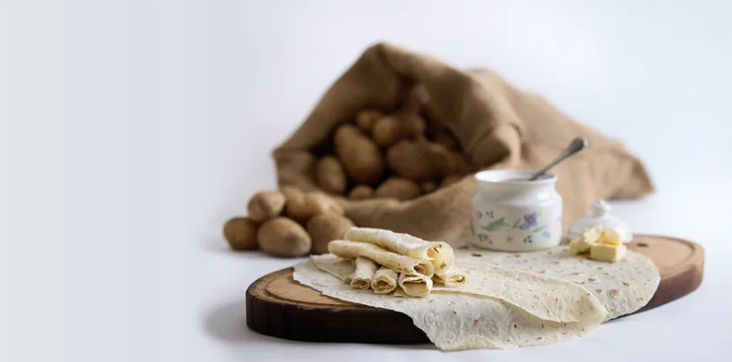 Flatbread and butter on a wooden board, with a sack of potatoes and a jar of cream in the background.