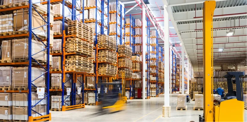 Wide warehouse aisle with tall blue-orange racks stacked with pallets and boxes; blurred yellow forklift moving past.