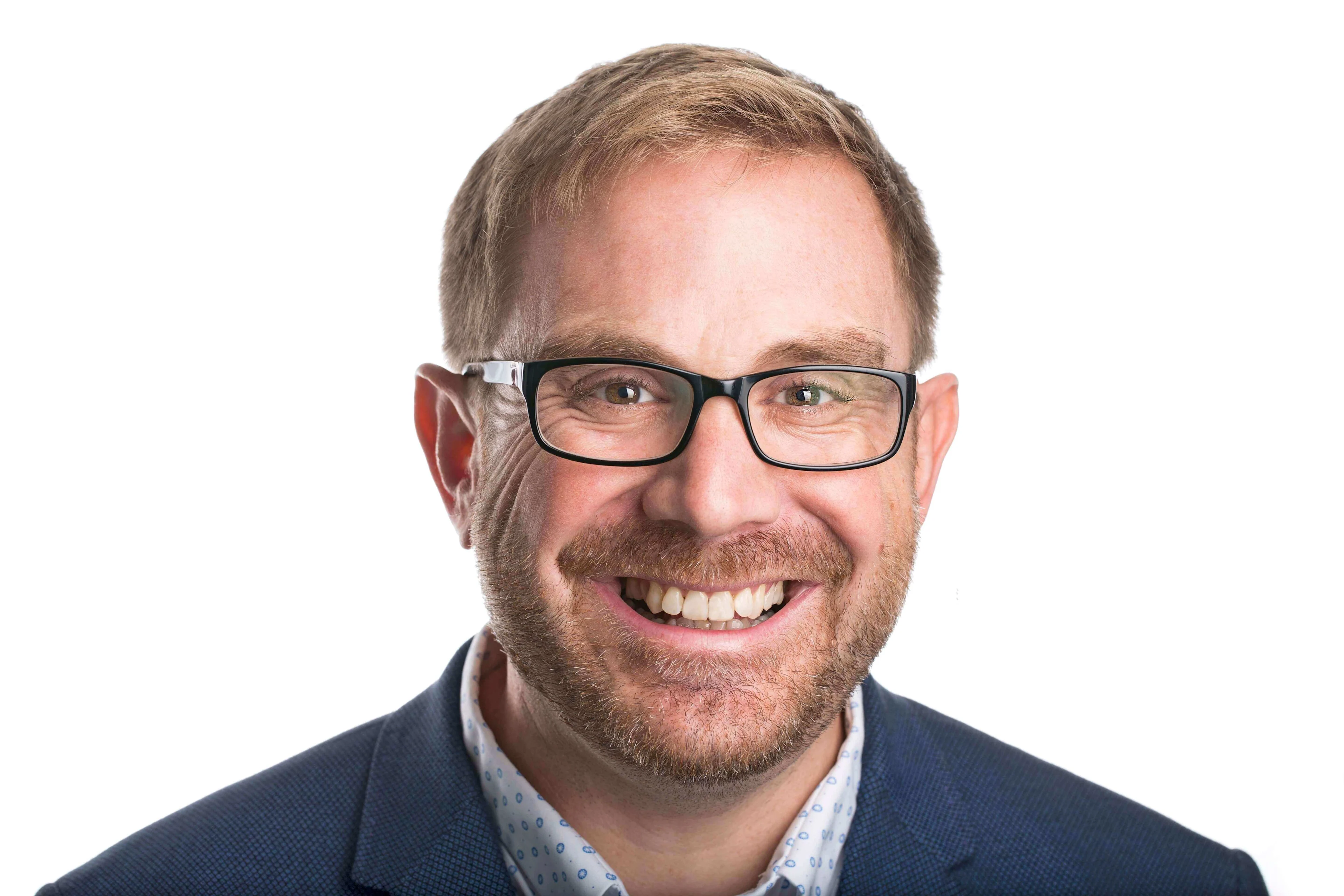Smiling man with light brown hair, beard, and glasses wearing a blue jacket and patterned shirt on a white background.