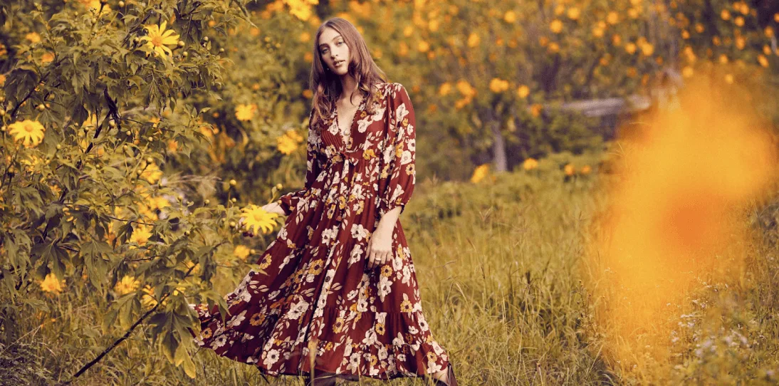 Woman wearing a flowing, dark floral print dress standing in a sunlit field of yellow flowers.