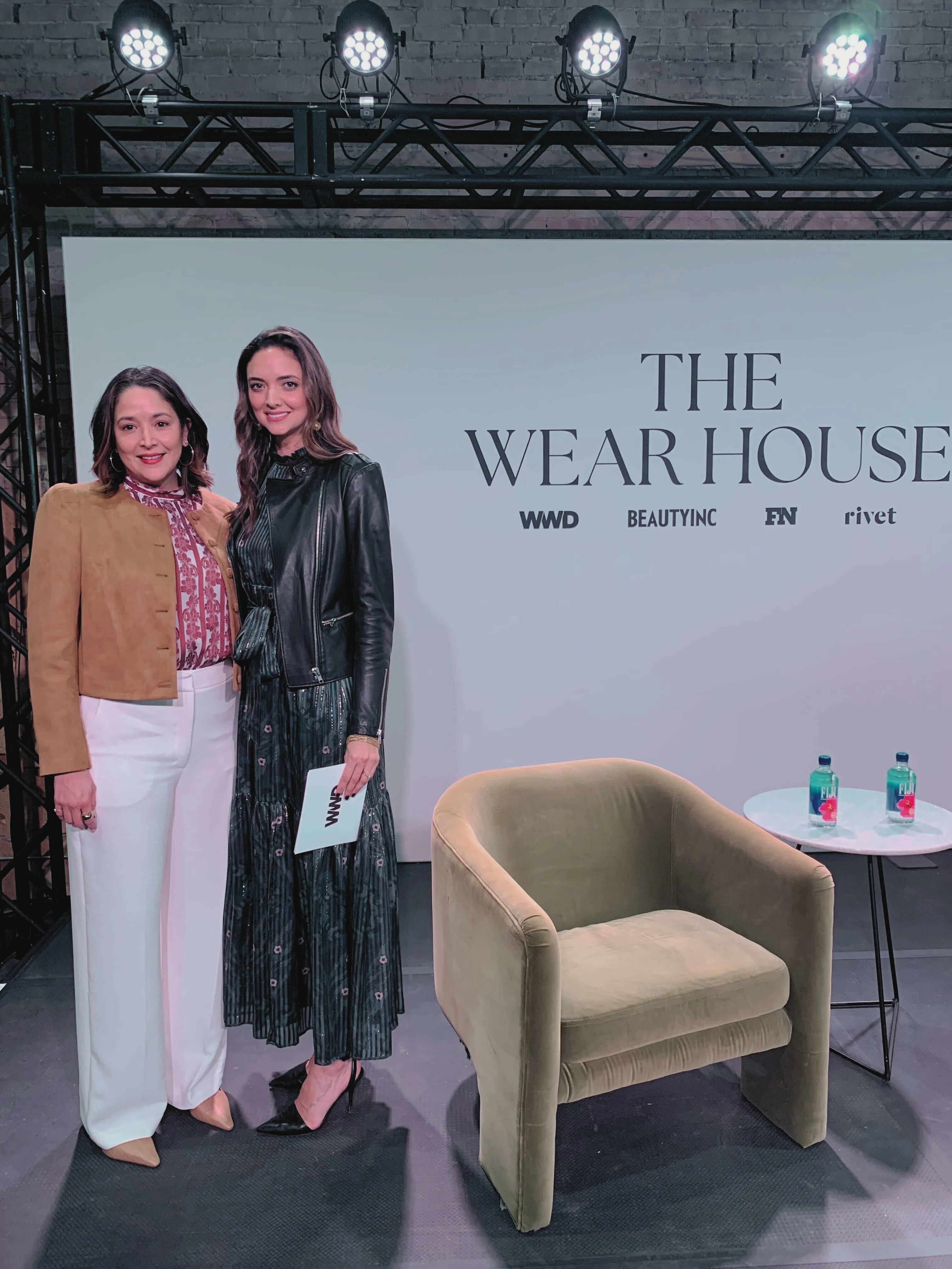 Two women pose on stage in front of a sign reading "The Wear House," beside a green chair and a small table with water bottles.
