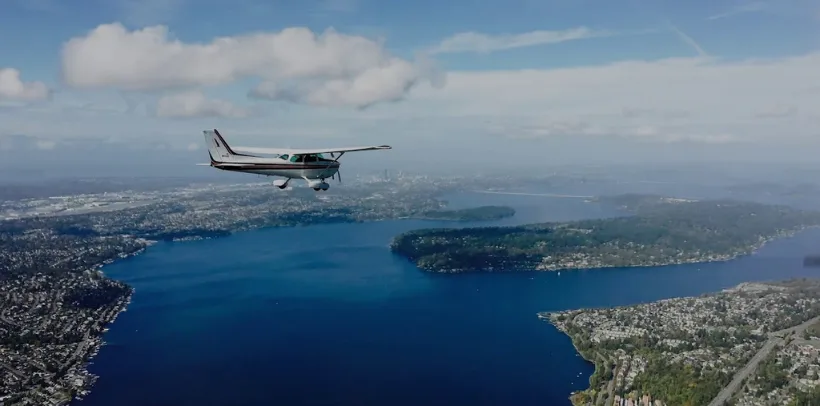 Small airplane flying over a large blue lake, surrounded by city and greenery, under a partly cloudy sky.