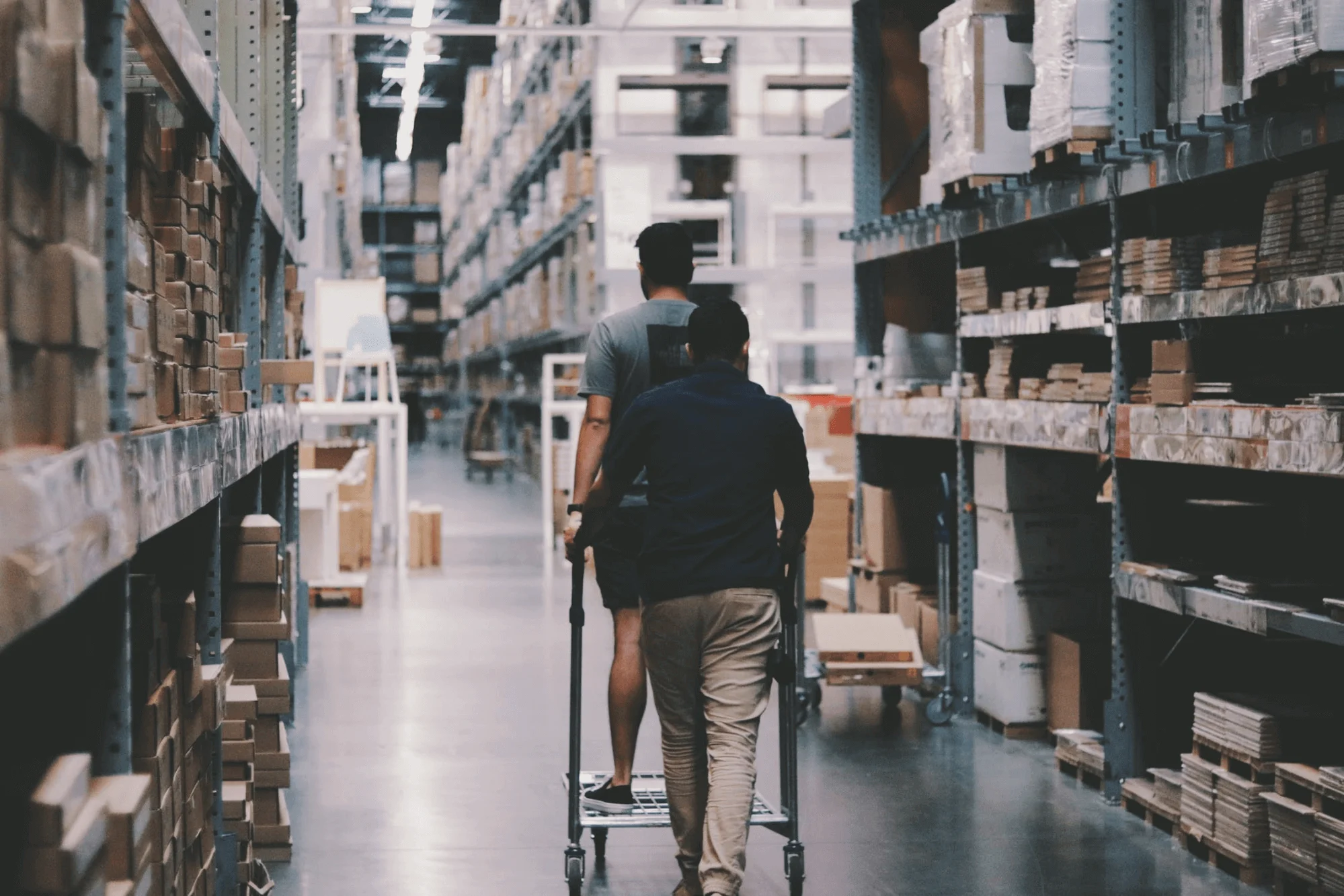 Two people in a warehouse, one pushing a cart with another person sitting, surrounded by shelves of boxes and packages.