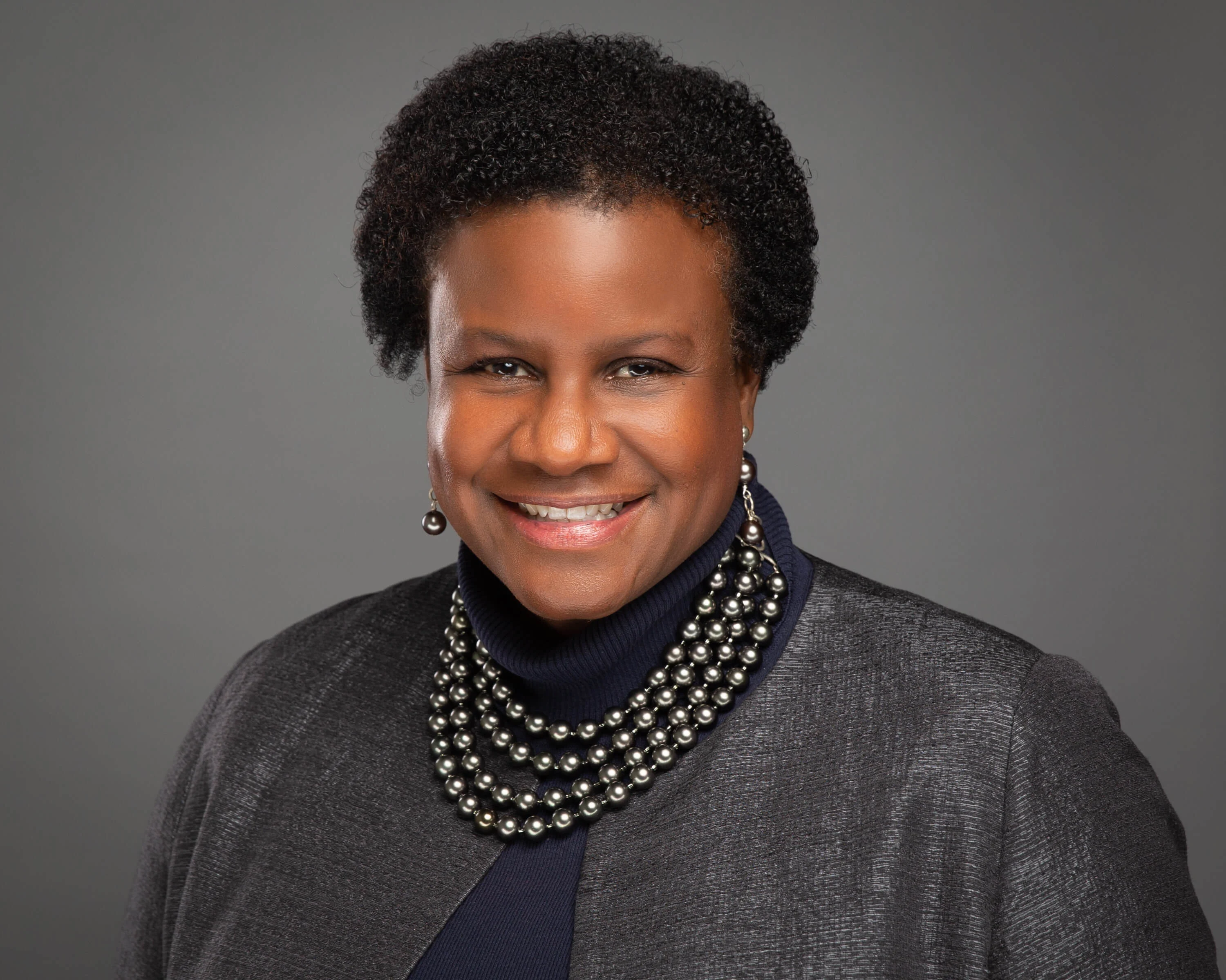 Smiling woman with short curly hair, wearing a dark jacket, layered pearl necklace, and earrings, against a gray background.