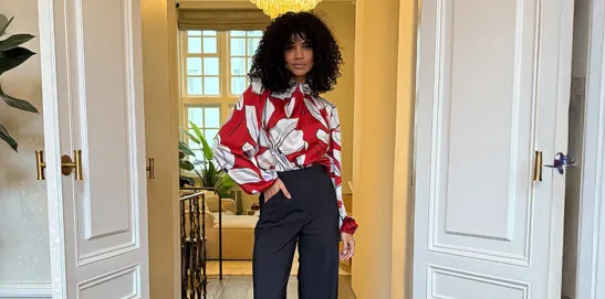 Woman with curly hair stands in a doorway wearing a red floral blouse and black pants, with a chandelier above.