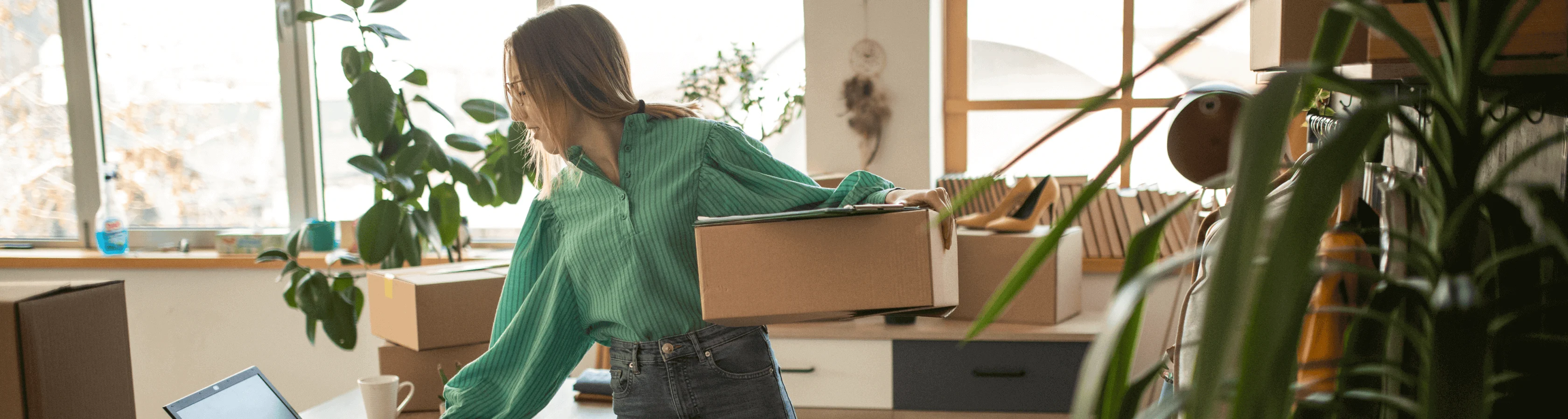 Woman in a bright office or home workspace holding a stack of cardboard boxes, ready to ship packages.