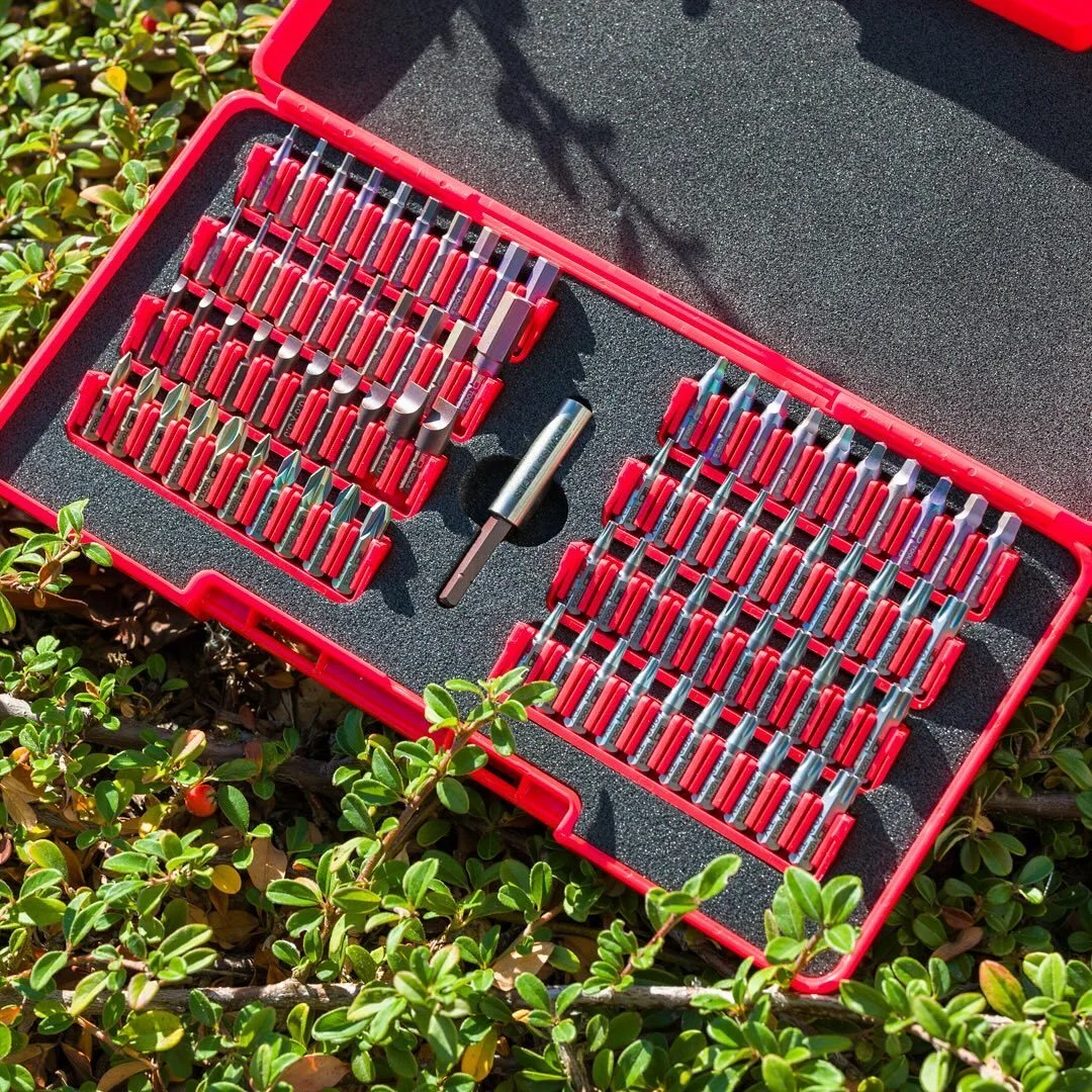 Red case with assorted screwdriver bits and a bit holder, open on green leafy ground in bright sunlight.