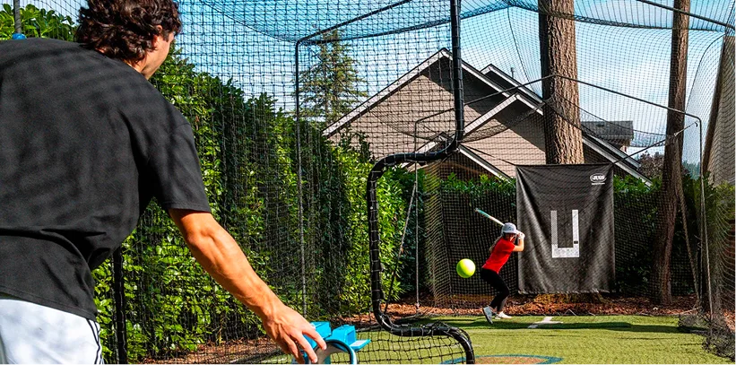 Person pitching baseball to batter in backyard batting cage with house and trees in background