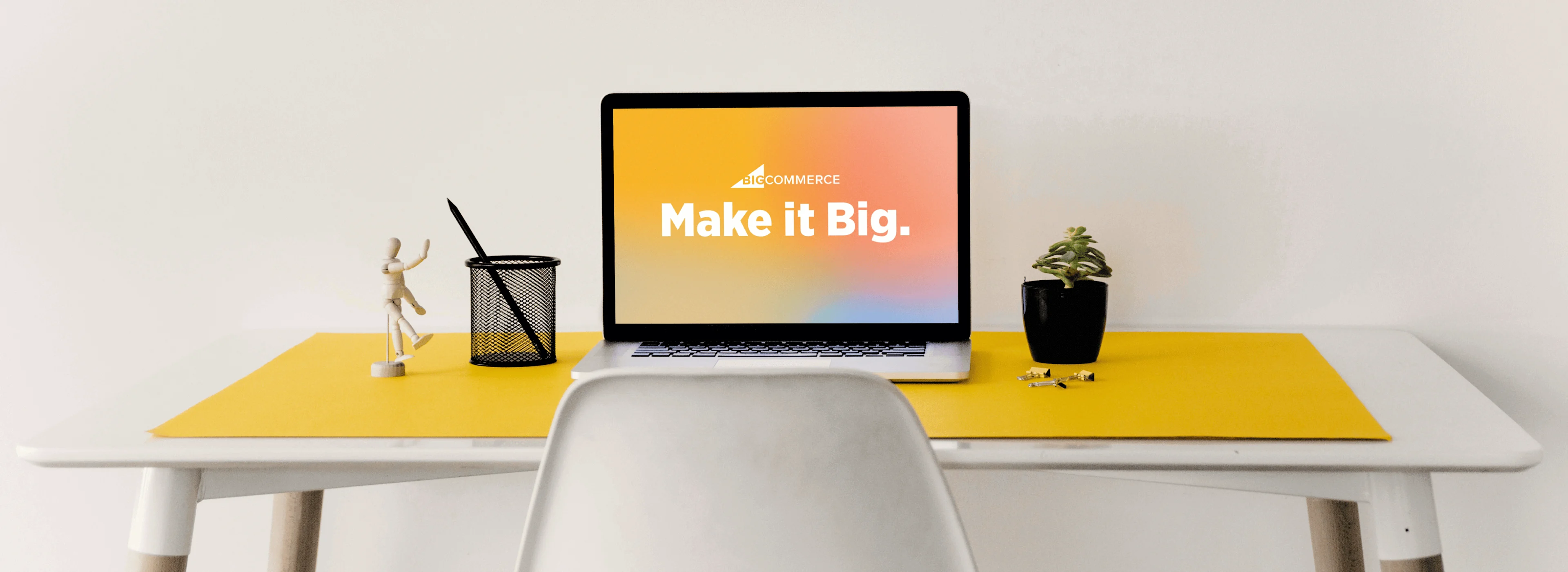 Minimalist desk with yellow mat, laptop displaying "Make it Big," pencil holder, wooden figure, and small potted plant.