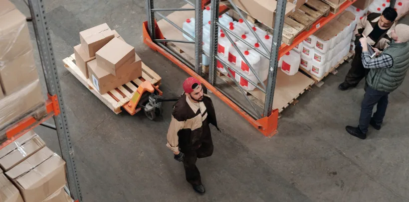 Warehouse worker pulls pallet of boxes while two people review documents near shelves with containers and packages.