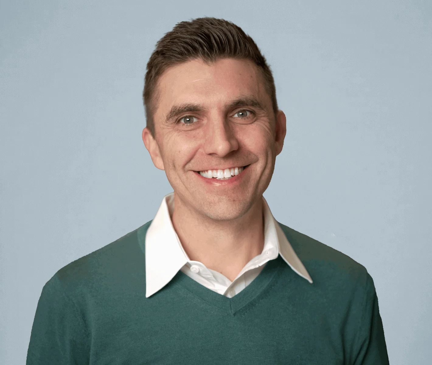 Smiling man with short brown hair wearing a green sweater over a white collared shirt, against a light blue background.