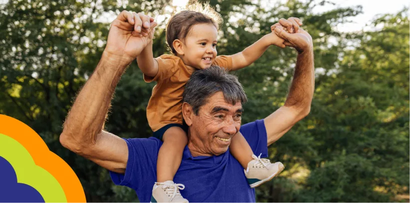 Smiling grandfather carrying young child on shoulders outdoors with green trees in background