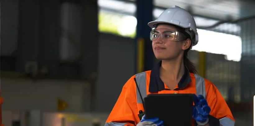 Woman in orange safety jacket, hard hat, and goggles holding a tablet in an industrial setting, looking to the side.
