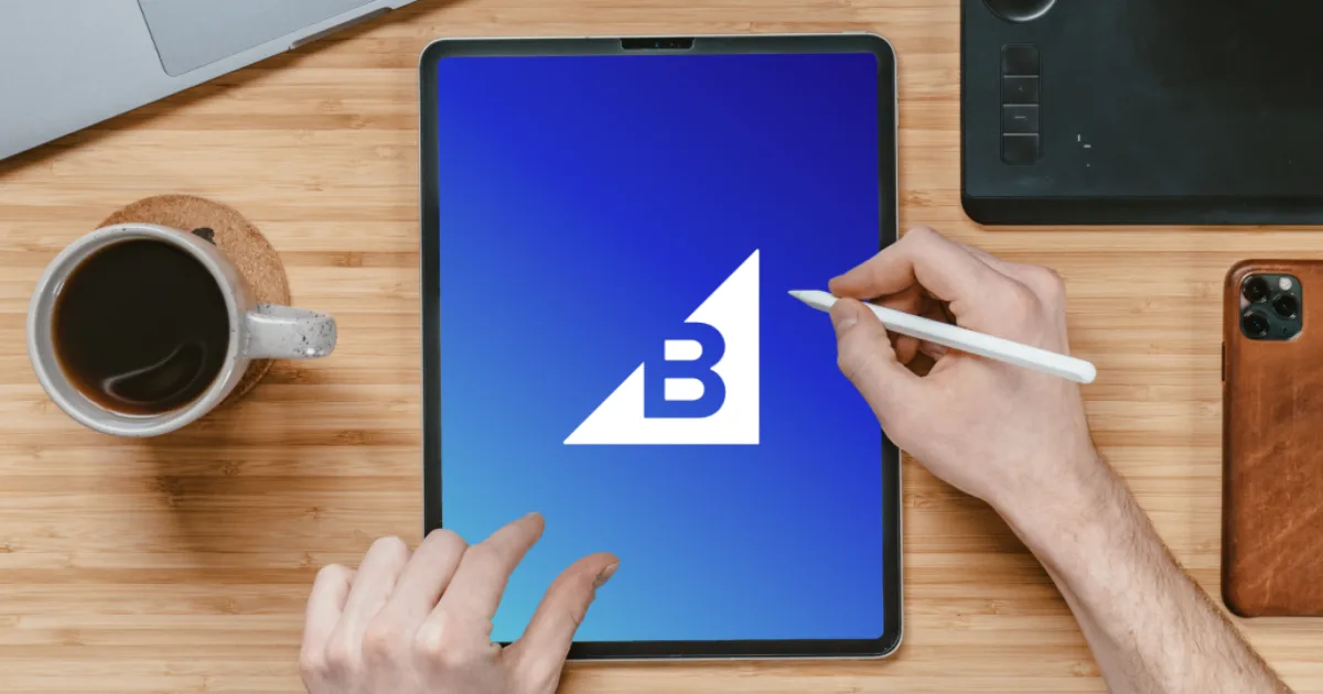 Person using a stylus on a tablet displaying a blue screen with a white B logo, coffee and devices nearby on a desk.