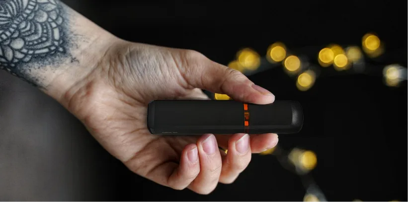Person holding a compact black vape device, with blurred lights in the background.