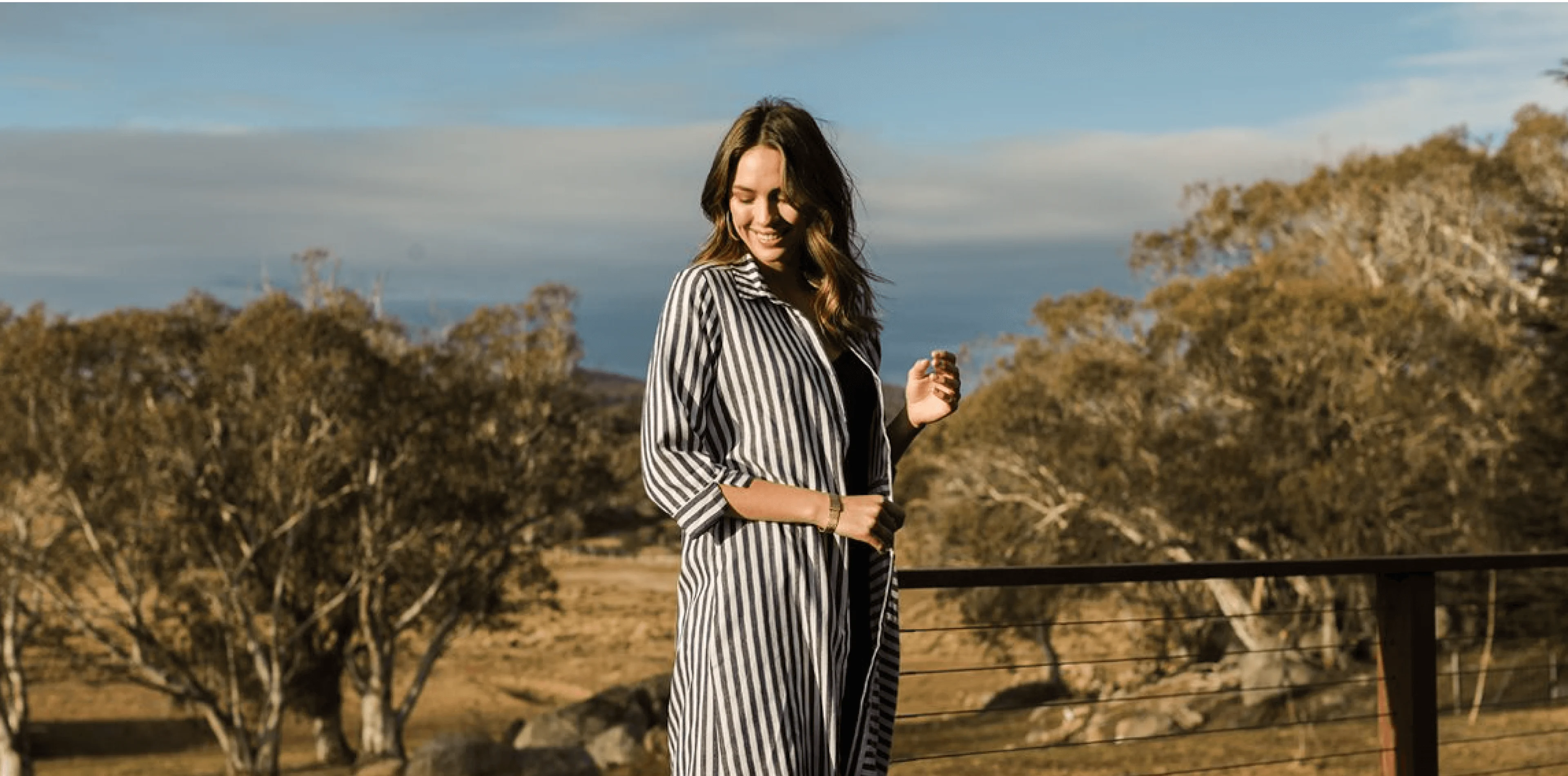 Woman in a black and white striped shirt smiling outdoors on a sunny day, with trees and blue sky in the background.