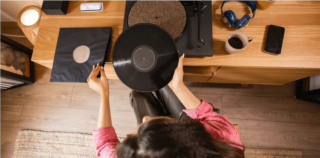 Overhead view of a woman holding a vinyl record, preparing to place it on a turntable on a wooden desk.