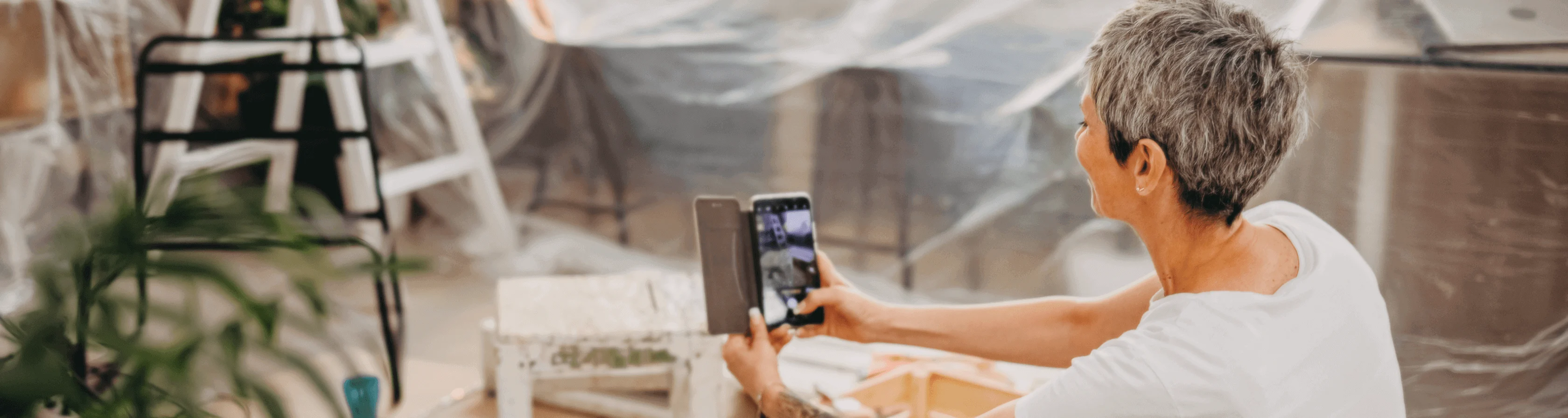 Smiling woman with short gray hair takes a photo on her phone in a room prepared for painting, with plastic sheets and plants.