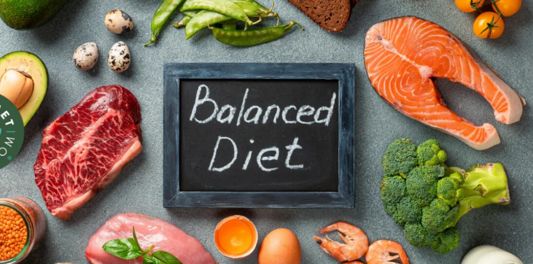 Assorted healthy foods like meat, fish, eggs, vegetables, and bread surround a chalkboard reading Balanced Diet.