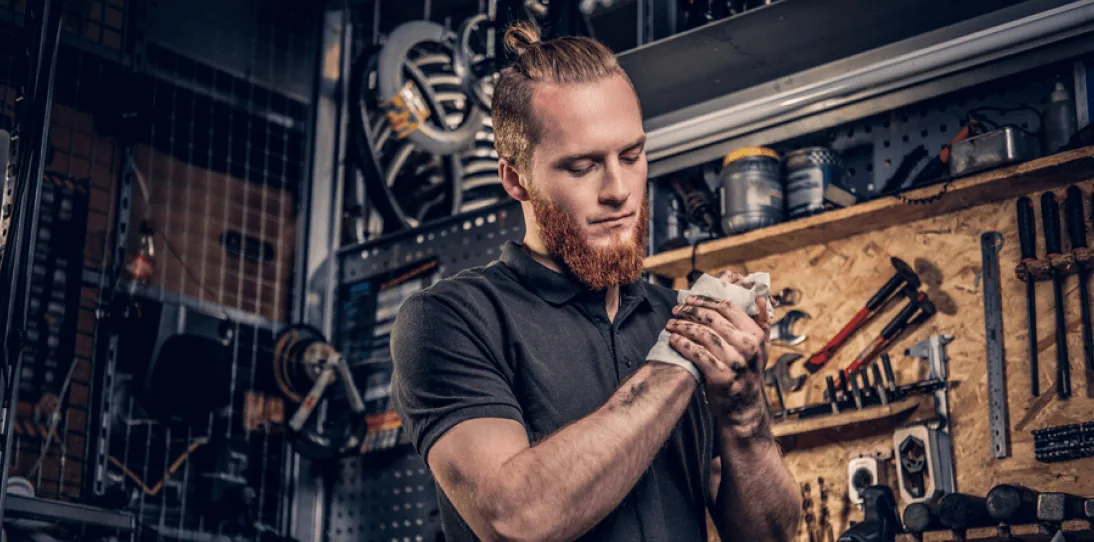 Man with a red beard wipes his hands with a cloth in a workshop filled with tools and equipment.