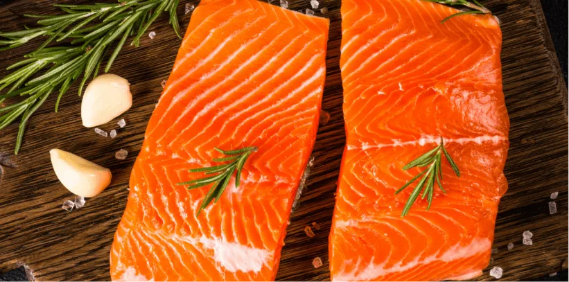 Two fresh salmon fillets with rosemary sprigs on a wooden cutting board, accompanied by garlic cloves and peppercorns.
