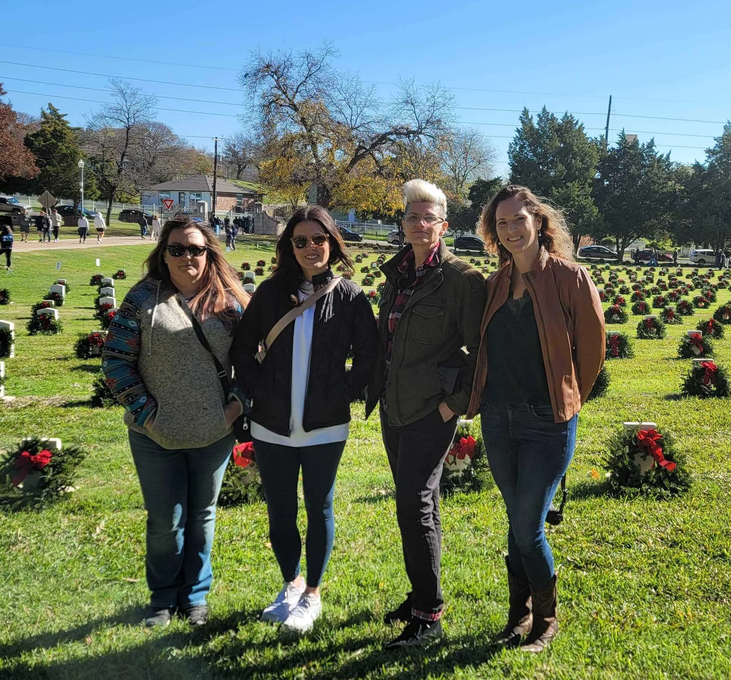 Four volunteers stand in a cemetery field adorned with wreaths on graves, during a sunny day.