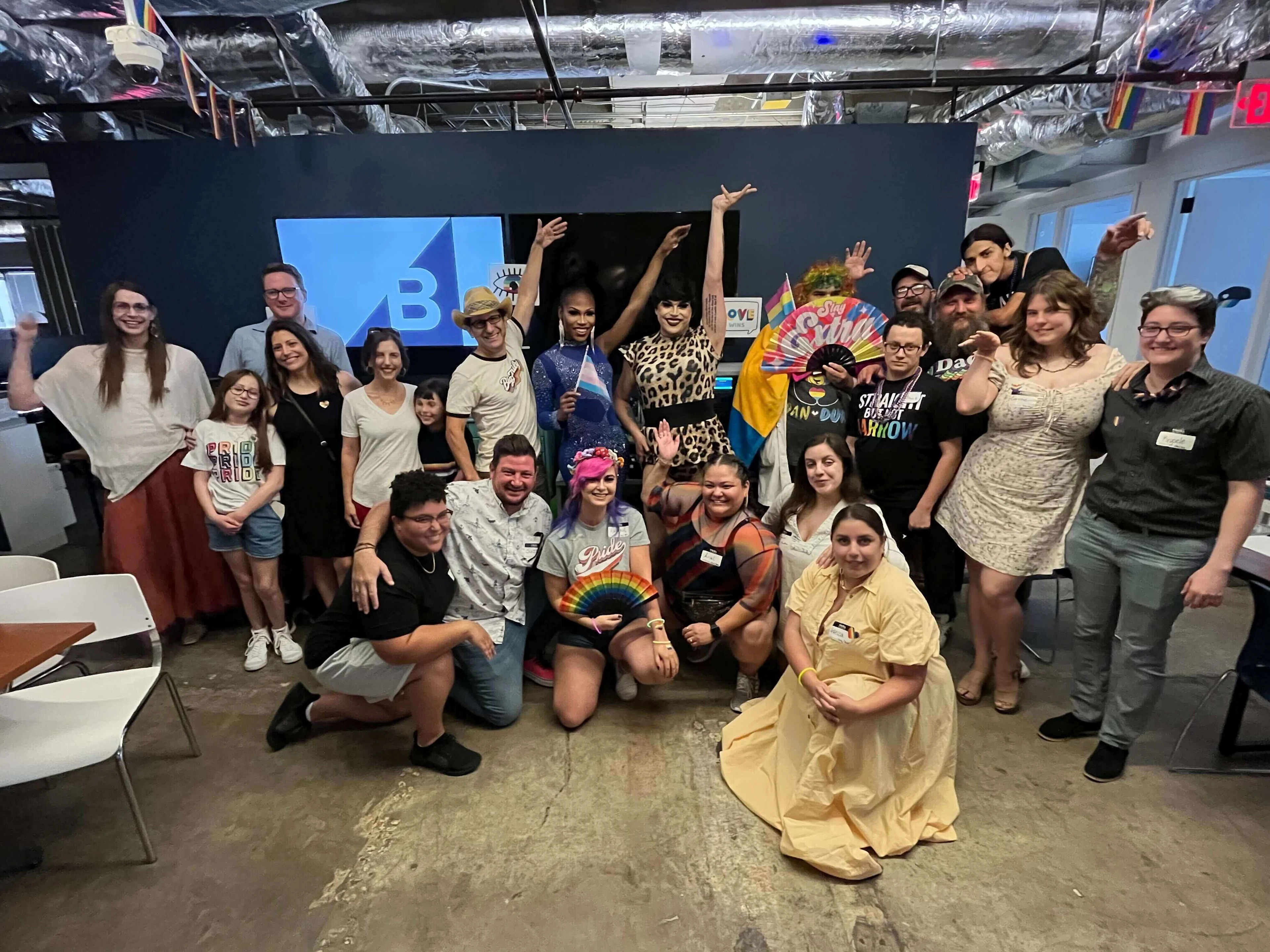 A large group of diverse people, some in drag and wearing Pride colors, pose together indoors, celebrating Austin Pride 2022.
