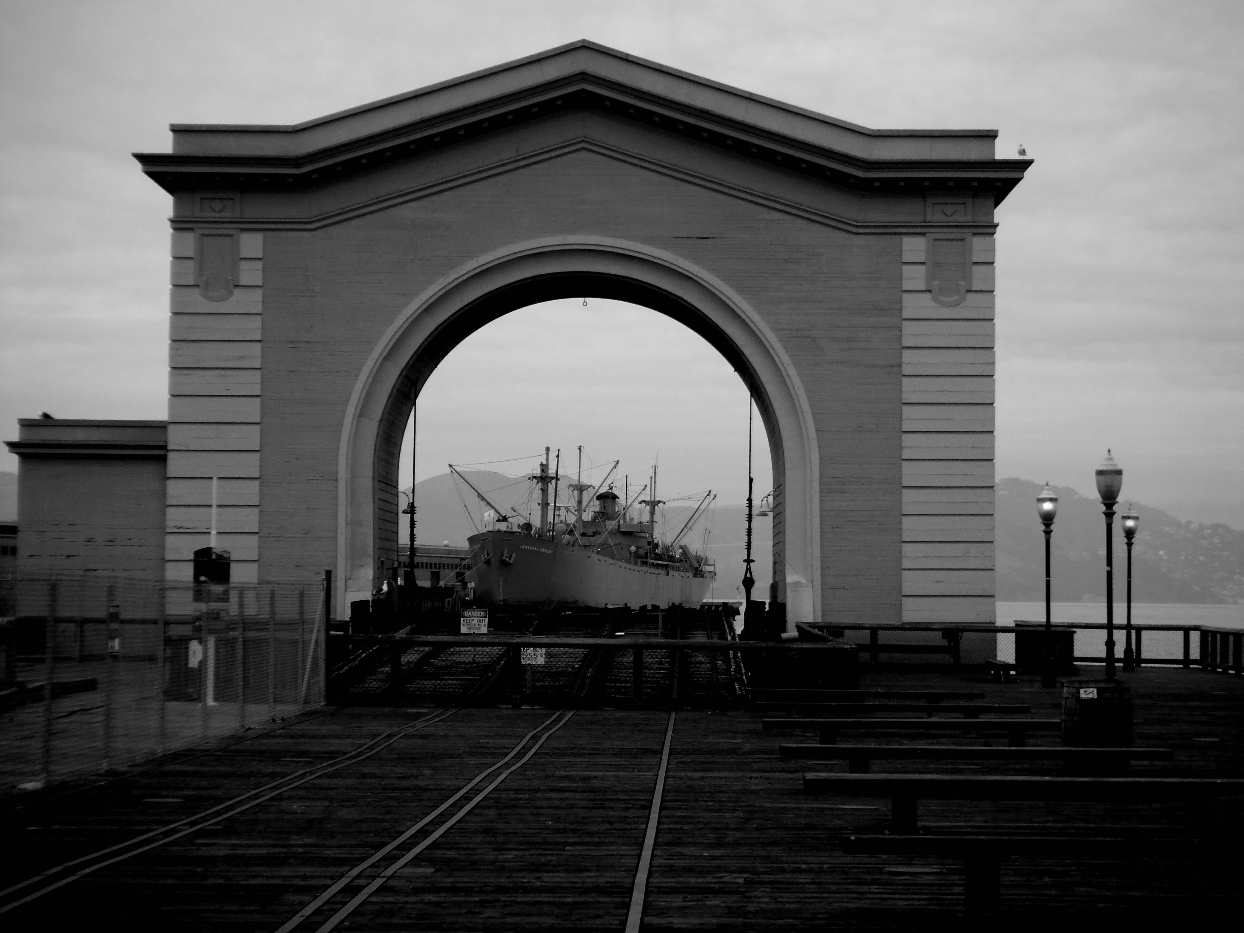 Archway leading to a boat resting in the water against a mountain background in black and white in Embarcadero, San Francisco, California. 