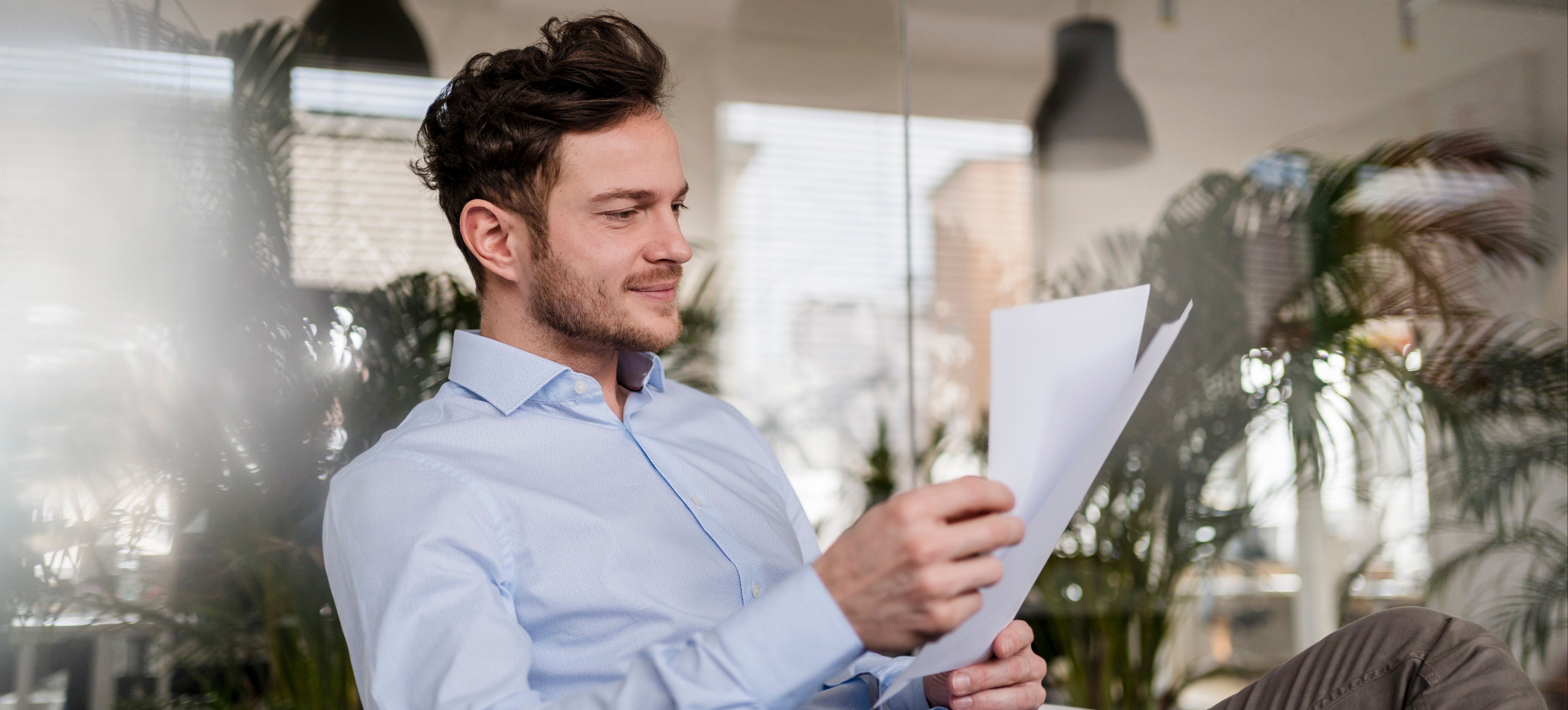 [Featured Image] A person in a light blue button-up is sitting down, holding and thoughtfully looking at two pieces of paper. 