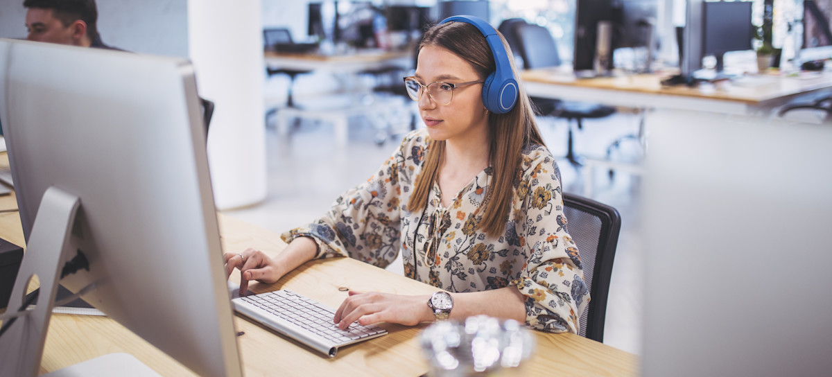 [Featured Image] A person wearing headphones works at a desktop on programming in Swift in an office. 
