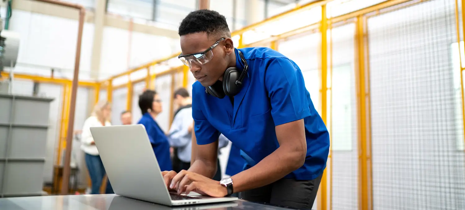 [Featured Image] A person working for a network operations engineer salary in an industry setting bends over their laptop.