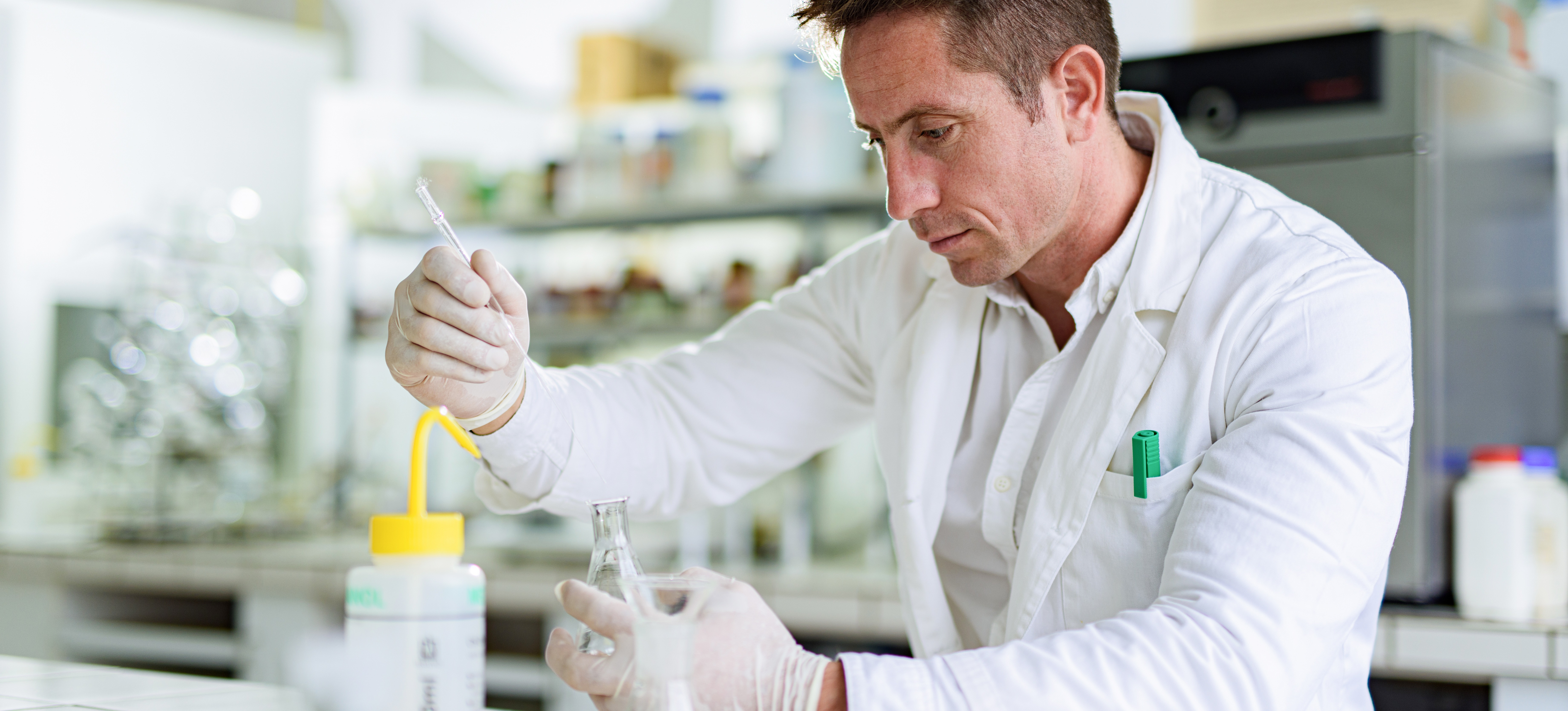 [Featured Image] A toxicologist wearing a white lab coat and white gloves carefully adds liquid to a beaker using a glass syringe while sitting in a lab.