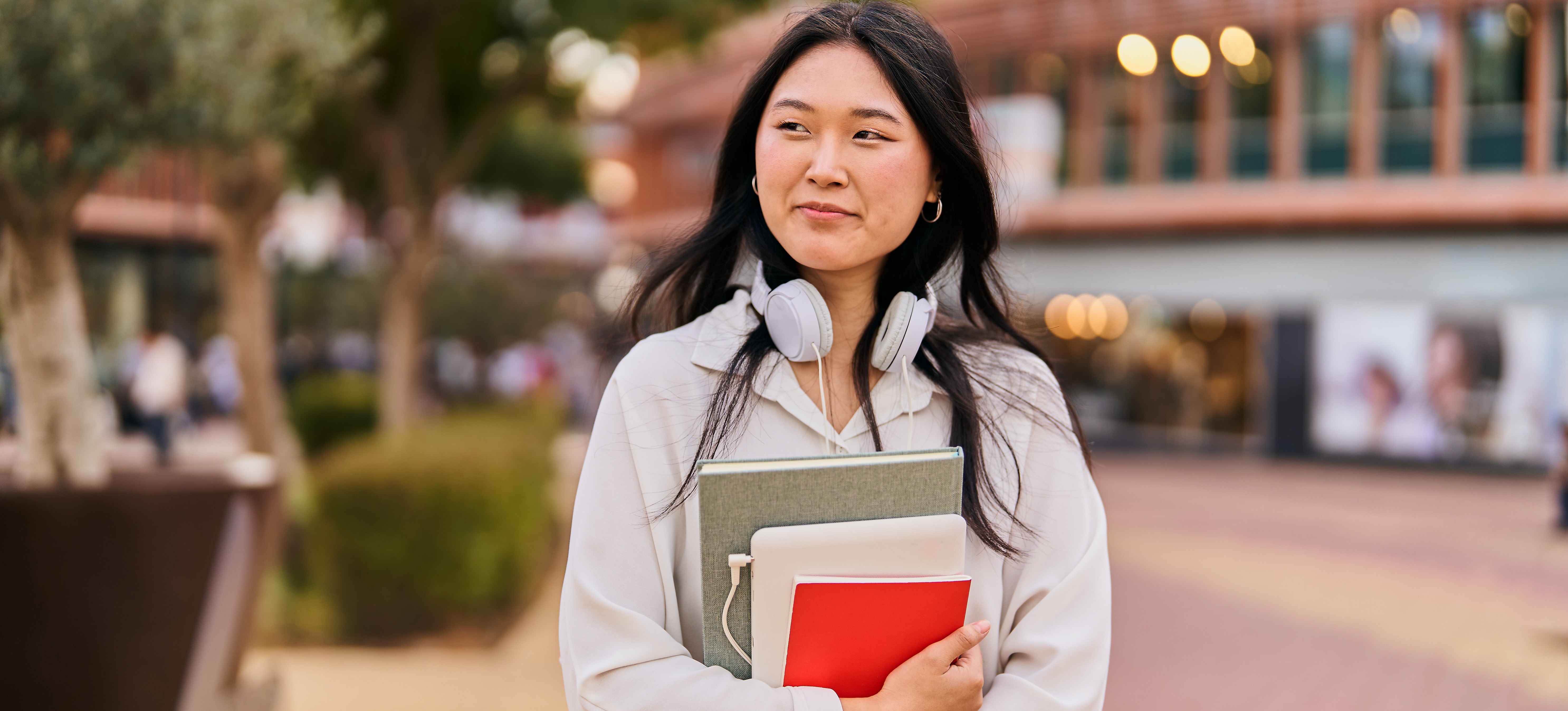 [Featured Image] A smiling learner holding books and wearing headphones around their neck walks on campus after getting into uni with no A levels.