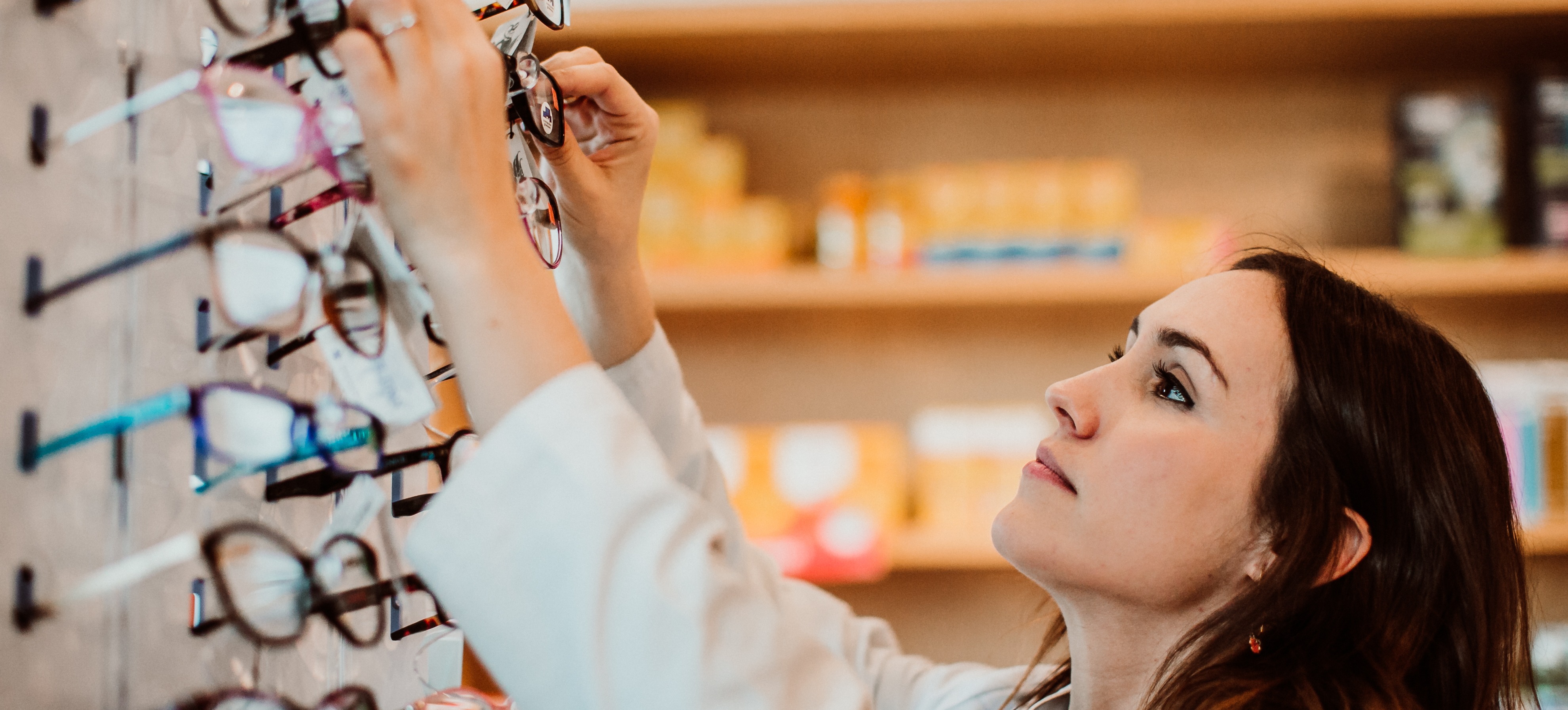 [Featured Image] An optician reaching up to select a pair of glasses from a selection of many, preparing them for a patient.

